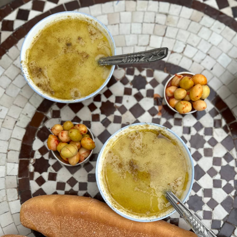 Two bowls of Moroccan soup with spoons, a small bowl of olives, and a baguette on a mosaic table during the Tangier food tour.