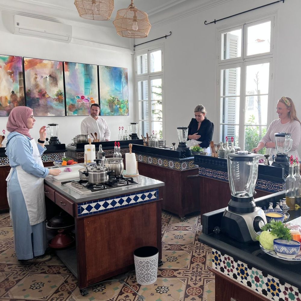 A cooking class with four participants in a well-lit kitchen. Each person is at a separate workstation with various appliances and ingredients, reminiscent of a Tangier food tour. The walls feature colorful artwork.