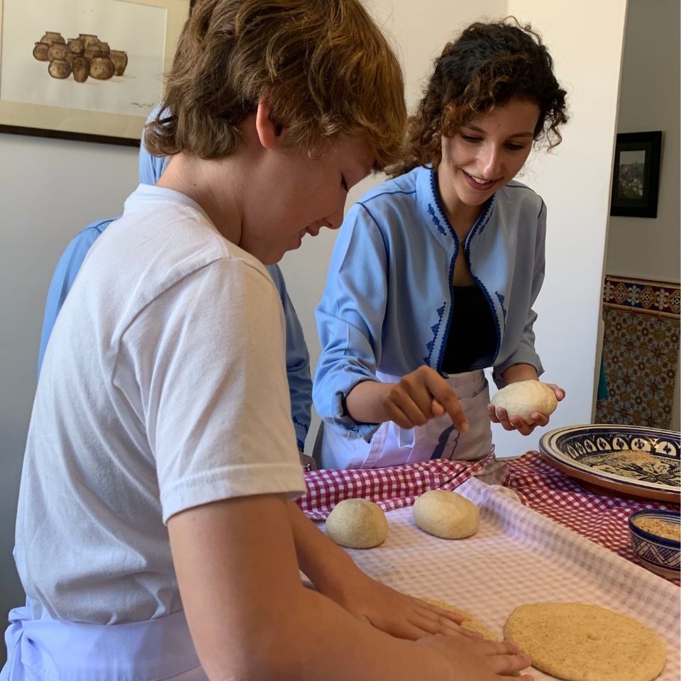 A young boy kneads dough while a woman instructs him. They are both smiling and working at a table covered with a red checkered cloth. More dough balls and a decorative plate are visible on the table.