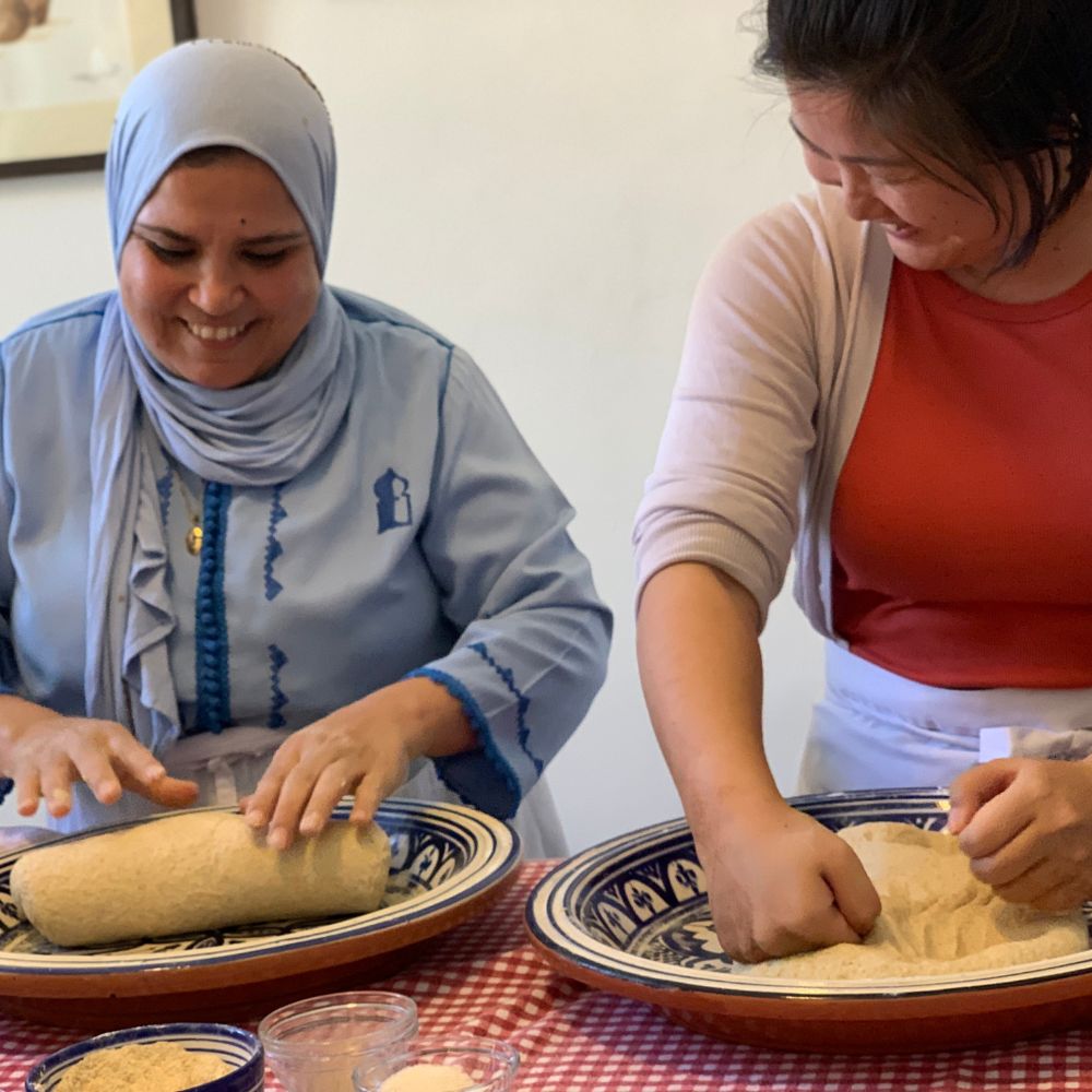 Two women, one in a blue hijab and the other in a red shirt, are smiling and working with dough in decorative bowls on a checkered tablecloth. Ingredients are spread across the table, capturing the vibrant atmosphere of a Tangier food tour.