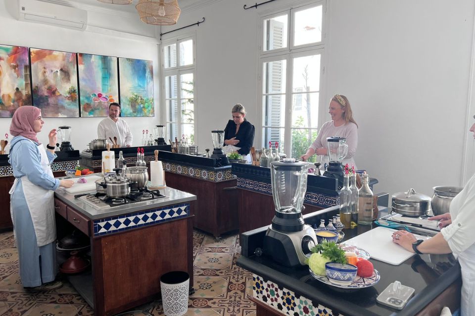 People participating in a cooking class in a bright kitchen. They are preparing ingredients and using cooking equipment on separate stations. Artwork is visible on the wall in the background.